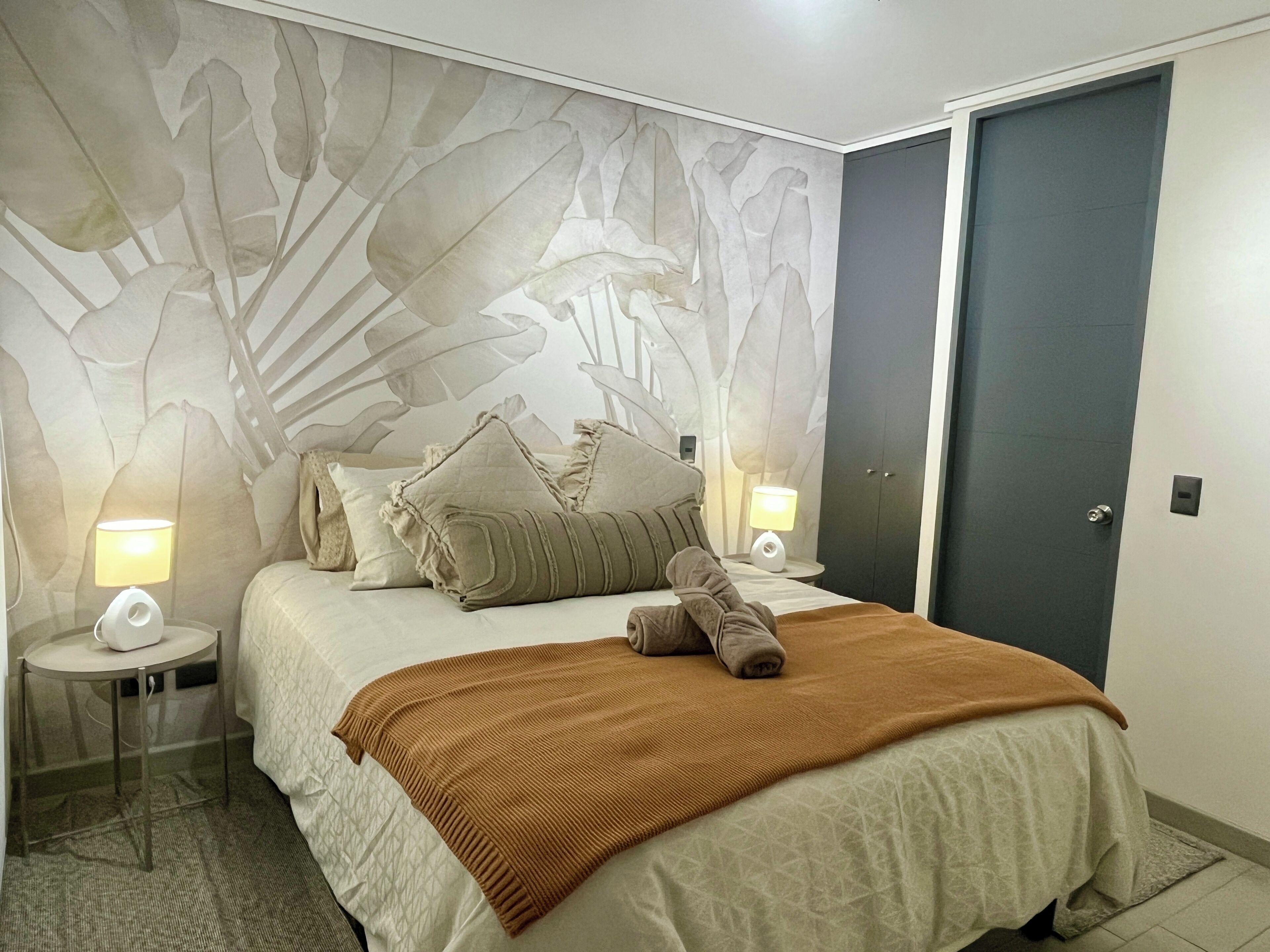 A hotel room with a bed, two bedside tables with lamps, a door, and a wall with a leaf pattern.