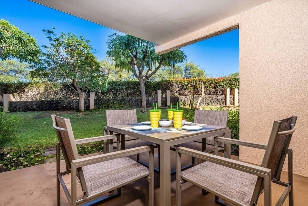A patio with a table set for four, surrounded by chairs, and a view of a garden with trees and a stone wall.