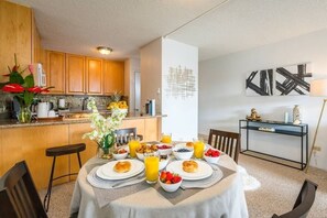 Une cuisine moderne avec des meubles en bois, une table à manger dressée pour le petit-déjeuner avec des fruits et des boissons, et une décoration minimaliste agrémentée d’œuvres d’art murales abstraites.