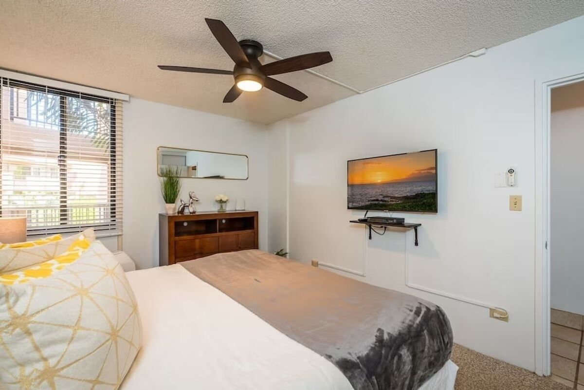 A bedroom with a bed, a ceiling fan, a TV on the wall, a dresser, and a window with blinds.