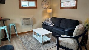 A living room with a dark sofa, a white coffee table, two bar stools, a lamp, and a clock on the wall.