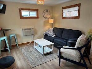 A living room with a dark sofa, a white coffee table, two bar stools, a lamp, and a clock on the wall.