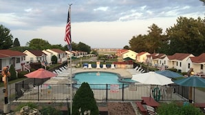 A swimming pool with lounge chairs and umbrellas, surrounded by a fence, with a flagpole and a view of houses in the background.