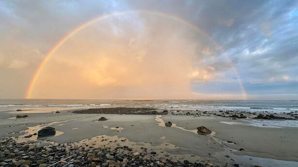 A beach with a rainbow, rocks, and a cloudy sky.