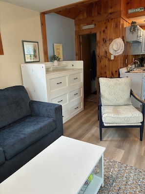 Una sala de estar con un sofá, un aparador blanco, un sillón estampado y una zona de cocina con paredes de madera y un letrero que dice 'Beach Bum Blvd'.