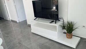 Una sala de estar moderna con un televisor de pantalla plana sobre un mueble blanco, un control remoto y una planta en maceta.