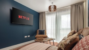 Una sala de estar moderna con un televisor de pantalla plana mostrando 'NETFLIX', un sofá cómodo, una mesita de centro con un juego de ajedrez y un amplio ventanal con cortinas.
