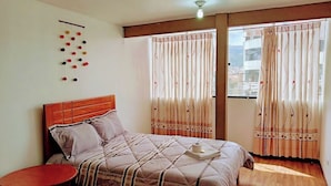 A bedroom with a wooden headboard, a bed with a striped quilt, a small round table, and curtains with a wavy pattern. - Samaya Department sharing (Cusco)
