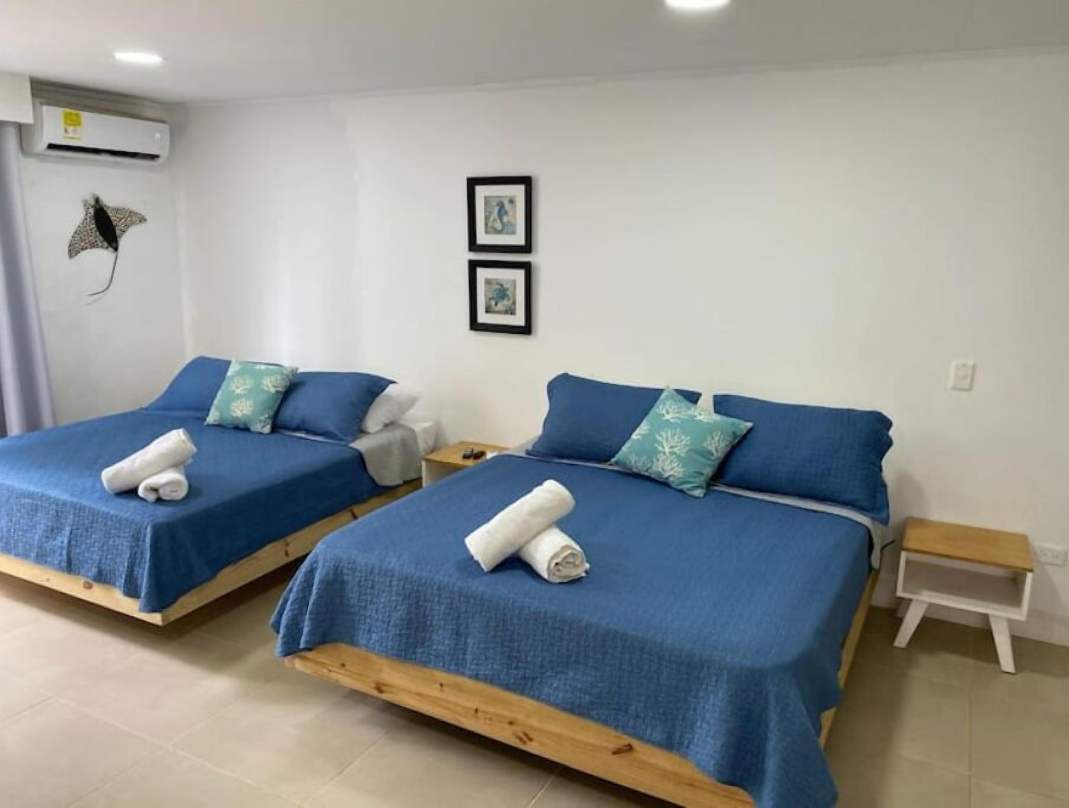 A room with two beds, each with blue bedding and decorative pillows. There is a small bedside table with a lamp on each bed.