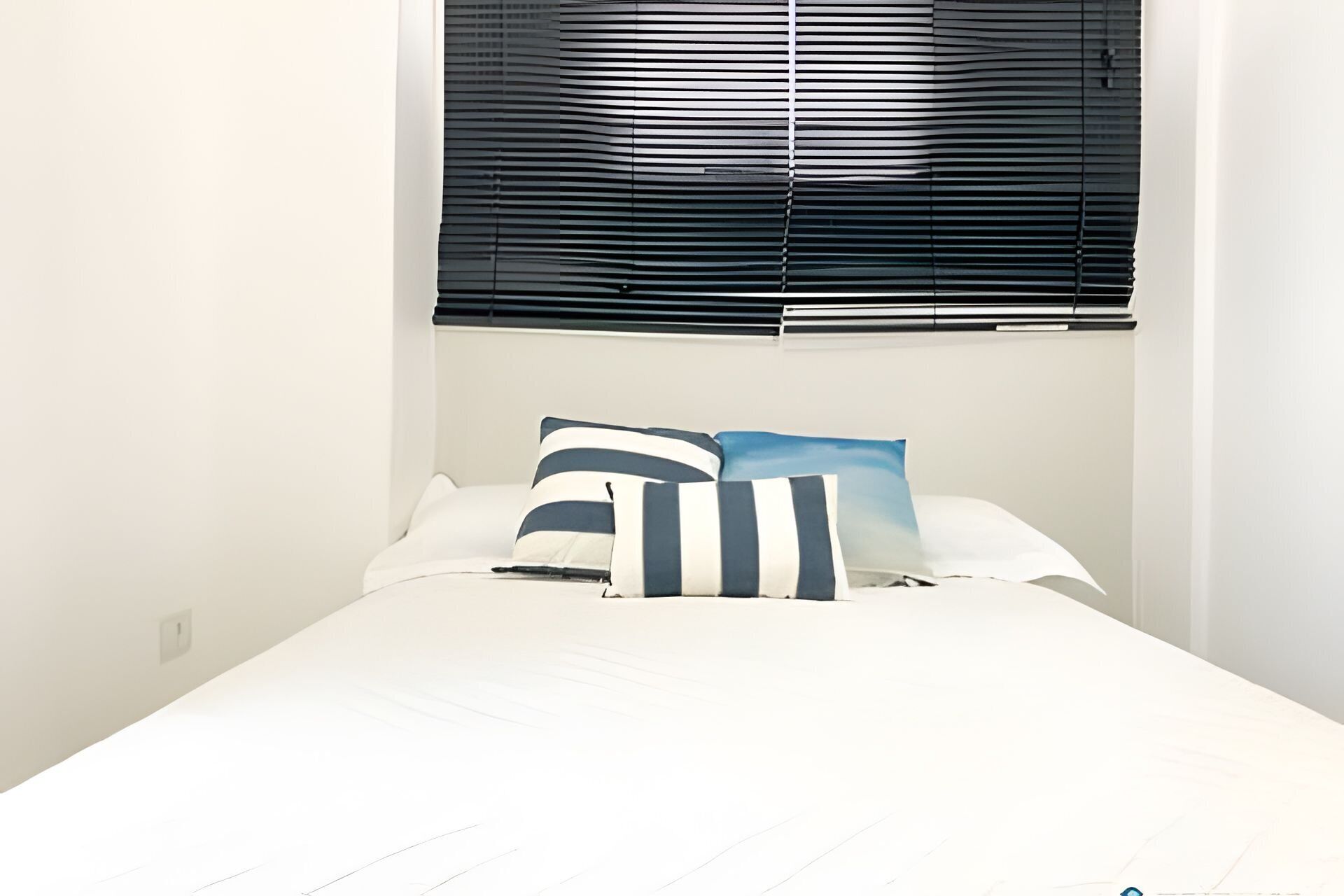 A bed with white bedding and two pillows, one striped and one solid blue, in a room with a window covered by horizontal blinds.