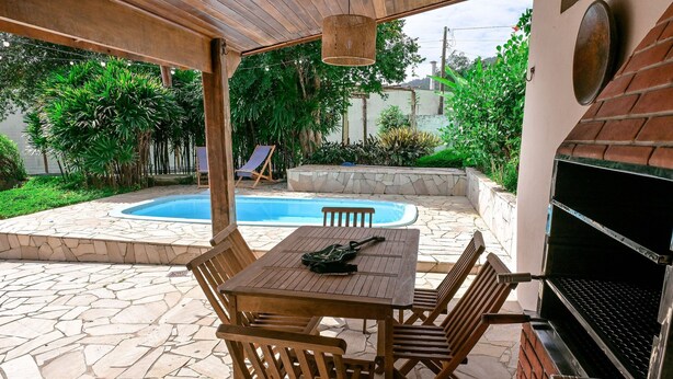 A patio with a stone floor, a wooden table with chairs, a grill, and a pool in the background.