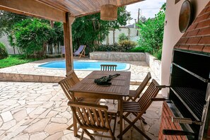 A patio with a stone floor, a wooden table with chairs, a grill, and a pool in the background.