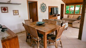 A dining area with a wooden table and wicker chairs, a television in the background, and a sofa in the adjacent living area.