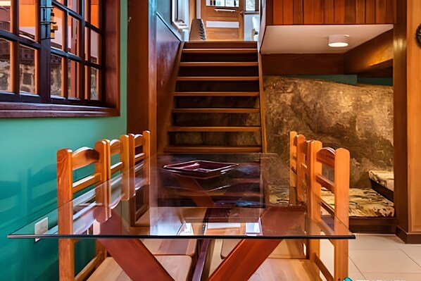 A dining area with a glass table and wooden chairs, a staircase, and a stone wall.