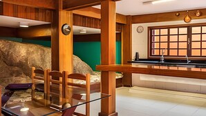 A kitchen with wooden beams, a stone wall, a dining area with chairs and a glass table, and a window with a view. - Birds' Home: Close to Curral Beach (Ilhabela)