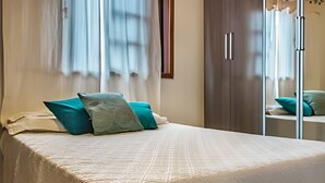 A neatly made bed with a white bedspread and two teal pillows, a wooden wardrobe, and a mirror reflecting the room's interior. - Birds' Home: Close to Curral Beach (Ilhabela)