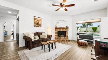 A living room with a fireplace, a ceiling fan, a large window, and a dining area.