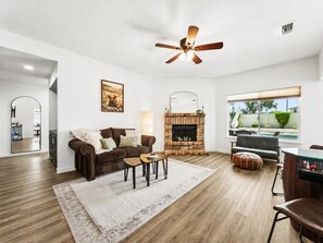 A living room with a fireplace, a ceiling fan, a large window, and a dining area.