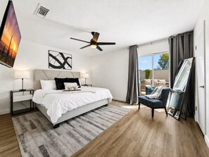 A modern bedroom with a bed, bedside tables, a ceiling fan, a large window with curtains, and a view of the outdoors.