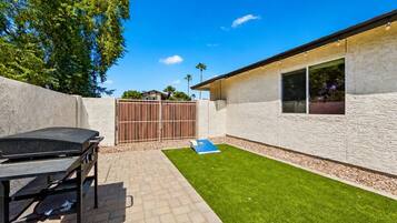 A backyard with a barbecue grill, a paved patio, a grassy area, and a building with a window.