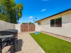 A backyard with a barbecue grill, a paved patio, a grassy area, and a building with a window.