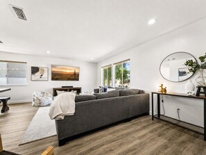 A modern living room with a grey sofa, a wooden floor, a dining table, and a large window.