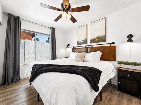 A bedroom with a bed, a ceiling fan, a nightstand, a window with curtains, and a wall-mounted lamp.