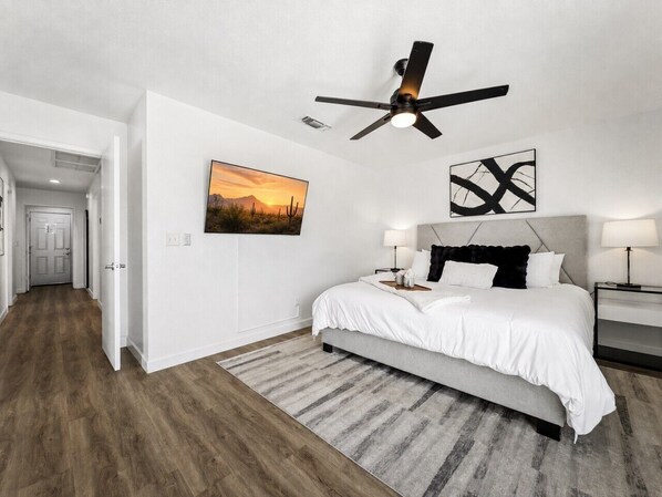 A bedroom with a bed, bedside tables, a ceiling fan, a wall-mounted artwork, and a door leading to another room.