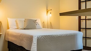A neatly made bed with a white bedspread and pillows, a bedside lamp, and a small round table. - Chalé da Libélula 3: Refuge close to the beach (Ilhabela)