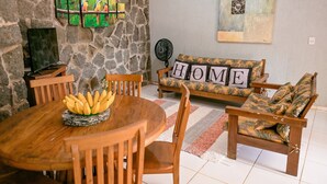 A dining area with a stone wall, a wooden table with a fruit bowl, a sofa with a 'HOME' sign, and a fan.