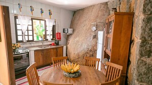 A kitchen with a round wooden table, chairs, and a fruit bowl. There is a stone wall, wooden cabinets, and a window with curtains.