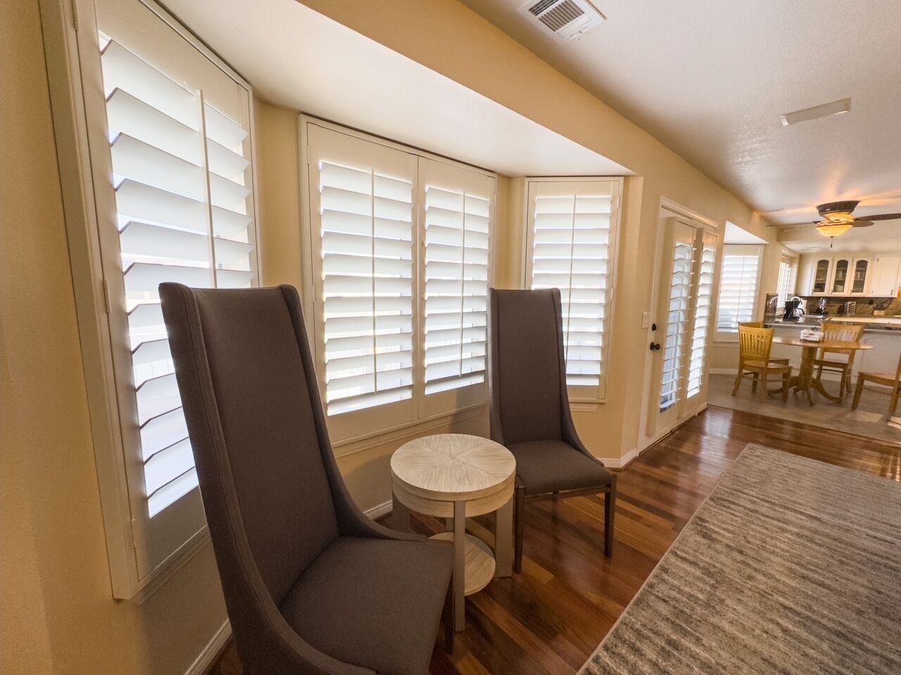 A spacious living room with two chairs and a small table, large windows with blinds, and a kitchen area in the background.