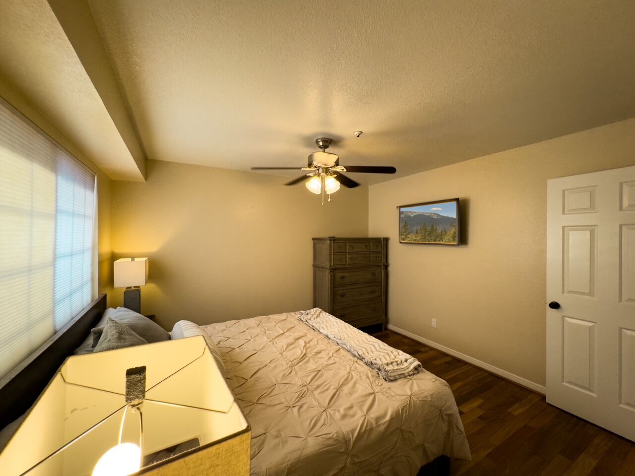 A bedroom with a bed, a ceiling fan, a dresser, a lamp, and a painting on the wall.