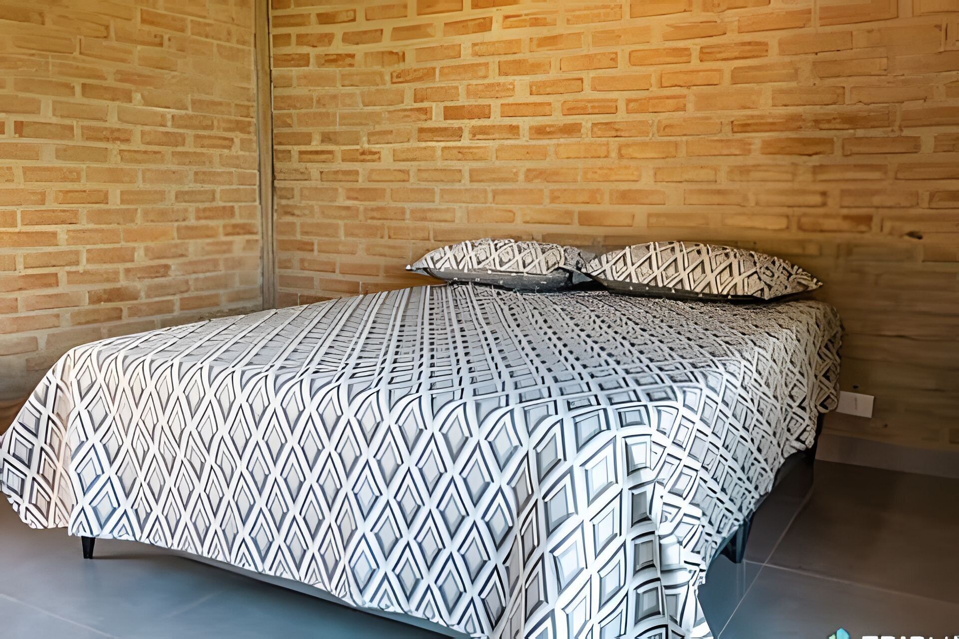 Una cama con una colcha de estampado geométrico y dos almohadas, frente a una pared de ladrillo.