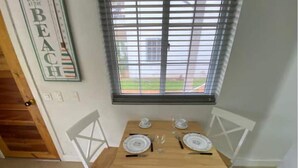 A dining area with a wooden table set for two, white chairs, and a window with blinds.