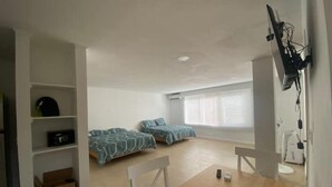 A small, bright room with two beds, a wooden table, and a TV mounted on the wall.