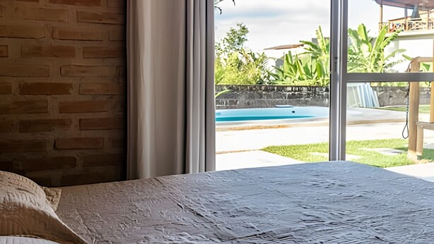 A room with a large window overlooking a swimming pool and greenery. - Recanto Feiticeira | Family suite in Feiticeira (Ilhabela)