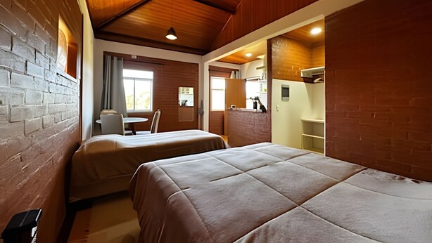 Una habitación con dos camas, una mesita pequeña y una silla. Hay una ventana con cortinas, una pared de ladrillo y un techo de madera.
