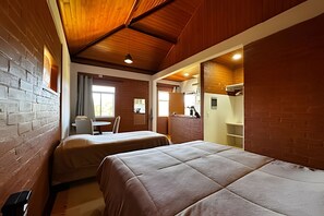 Une pièce avec deux lits, une petite table et une chaise. Il y a une fenêtre avec des rideaux, un mur en briques et un plafond en bois.