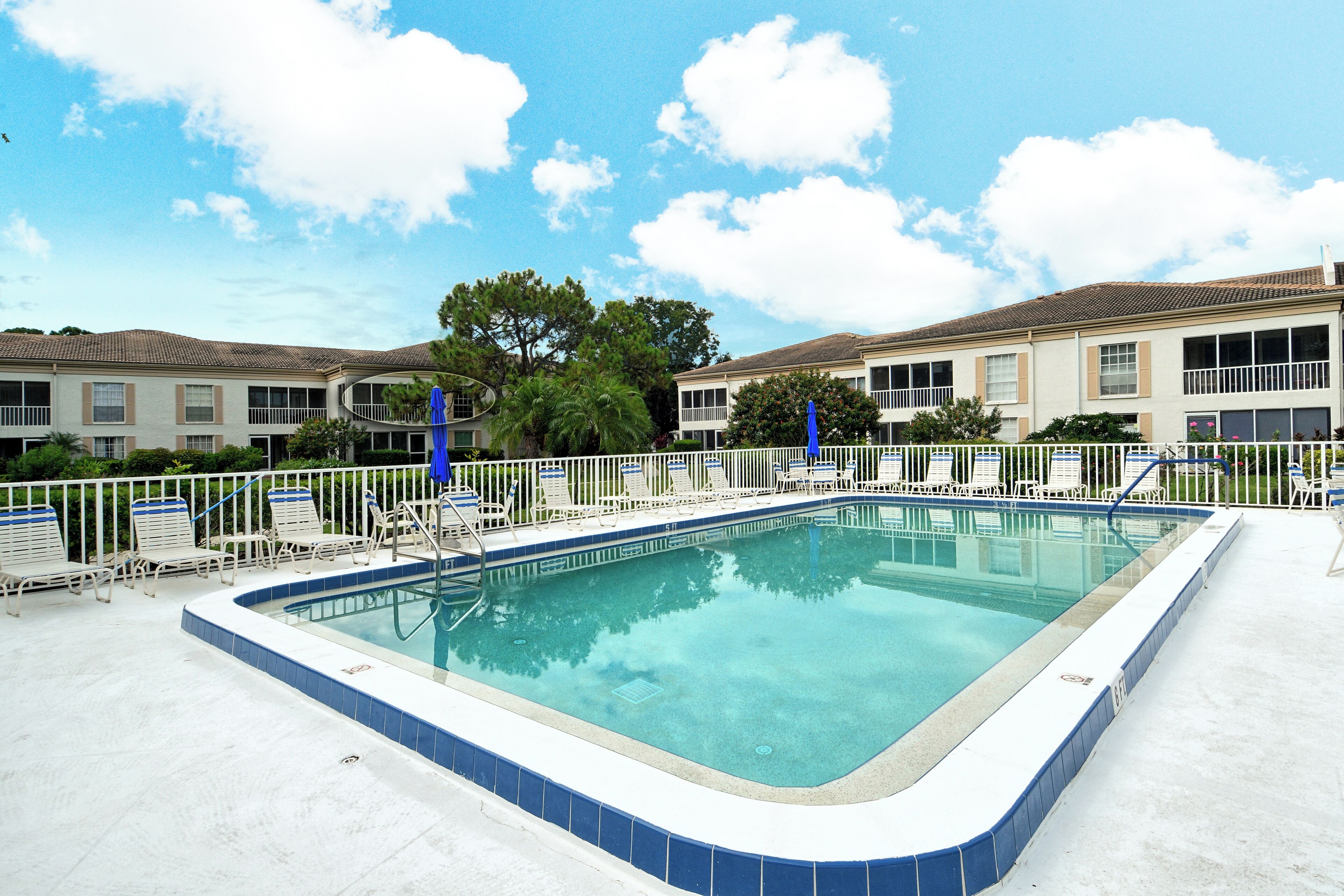 Una piscina rectangular con borde de azulejos azules, rodeada de sillones y sombrillas blancas, y un edificio al fondo.