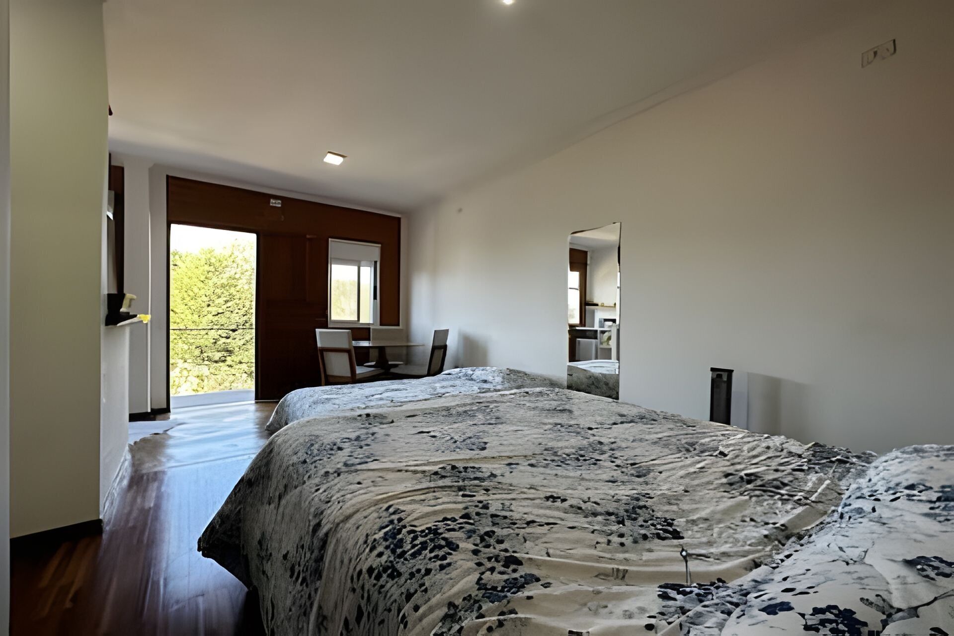 Ein Schlafzimmer mit einem großen Bett, einem Schreibtisch und einem Stuhl. Eine Tür führt zu einem Balkon mit Blick auf Bäume.