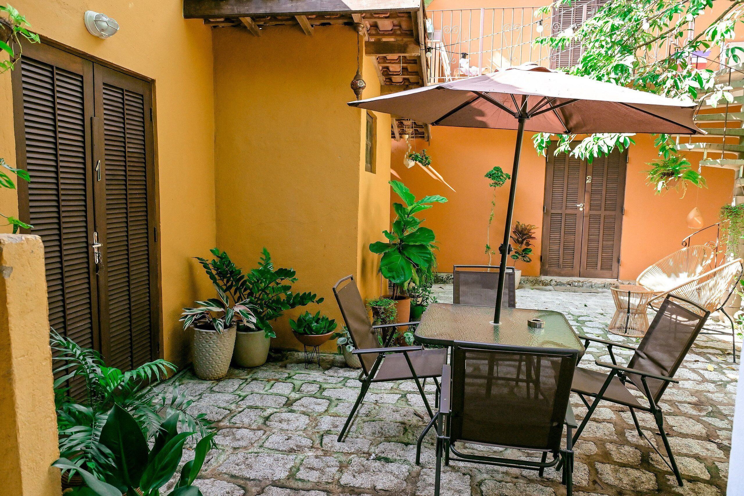 Une cour avec une table et des chaises placées sous un grand parasol, entourée de plantes en pot et dotée d’un sol en pierre.