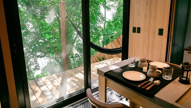 Un coin repas avec une table dressée pour deux personnes, une vue sur une terrasse en bois et une grande fenêtre donnant sur un espace verdoyant.