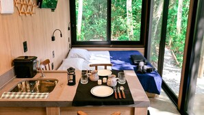 Une chambre moderne et compacte, dotée d’un lit, d’une petite kitchenette et d’une grande fenêtre donnant sur une forêt.