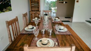 A dining table set with plates, glasses, and cutlery, with a wooden chair on one side. - Ponta do Sol | 4 suítes, piscina e churrasqueira (Ilhabela)