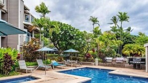 A swimming pool area with lounge chairs and umbrellas, surrounded by lush greenery and palm trees. - Grand Champions (Kihei)
