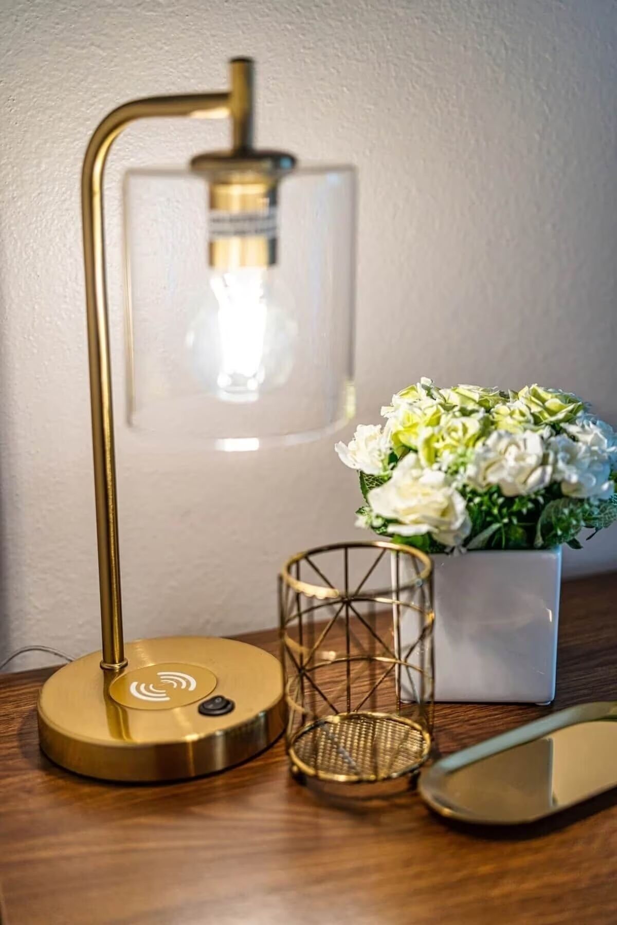 A desk lamp with a gold finish, a clear vase with white flowers, and a small wire basket.