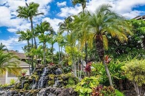 A tropical garden with a waterfall, palm trees, and a house. - Grand Champions (Kihei)