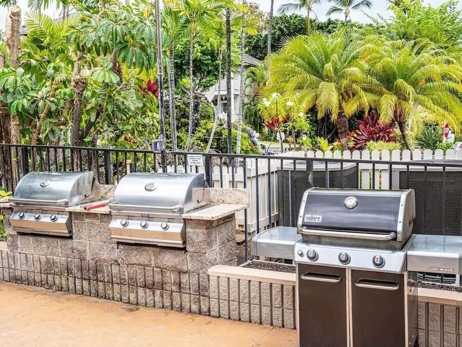 Uma área de churrasco com três grelhadores, uma parede de pedra e vegetação tropical exuberante ao fundo.