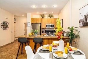 A modern kitchen with wooden cabinets, a stainless steel refrigerator, and a microwave. There is a dining table set with breakfast items and a vase of flowers. - Grand Champions (Kihei)